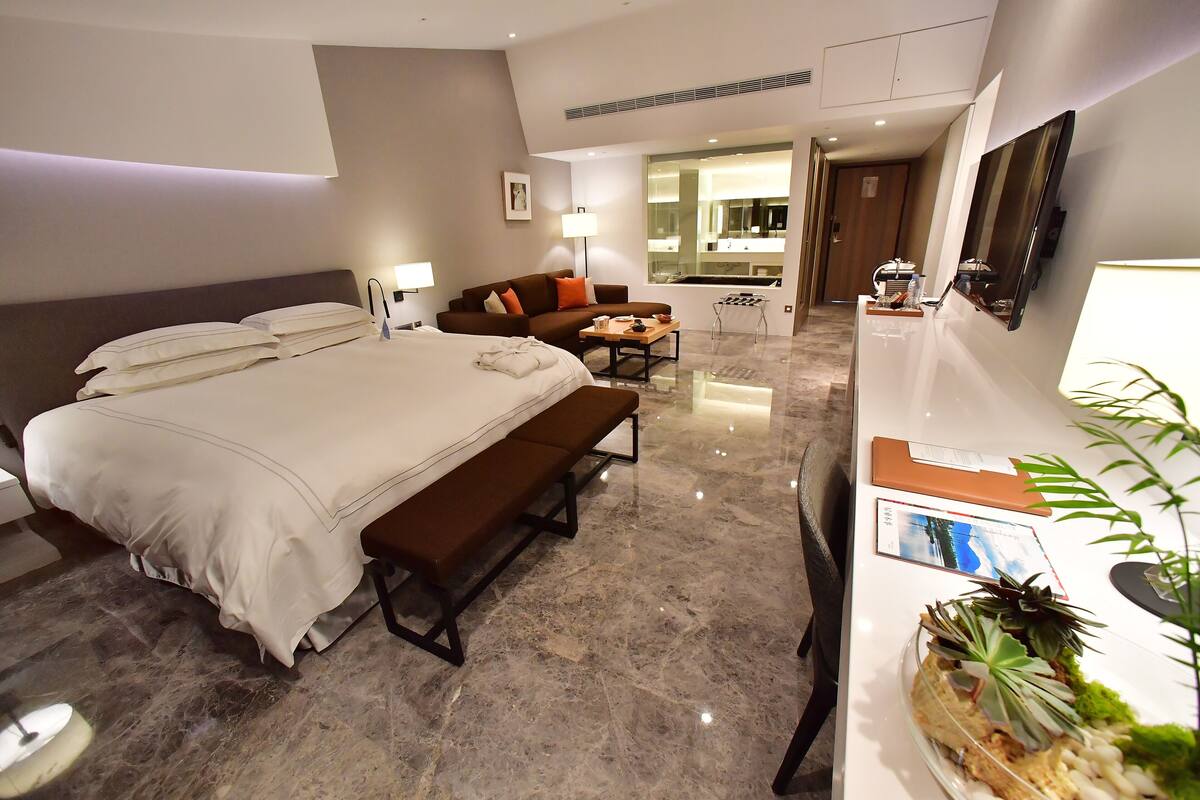 A hotel room with a bed, a bench, a desk, a mirror, and a plant.