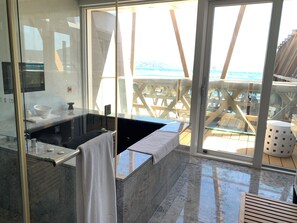Ein modernes Badezimmer mit einer großen Badewanne, einer Duschkabine aus Glas und Blick auf den Strand durch bodentiefe Fenster.