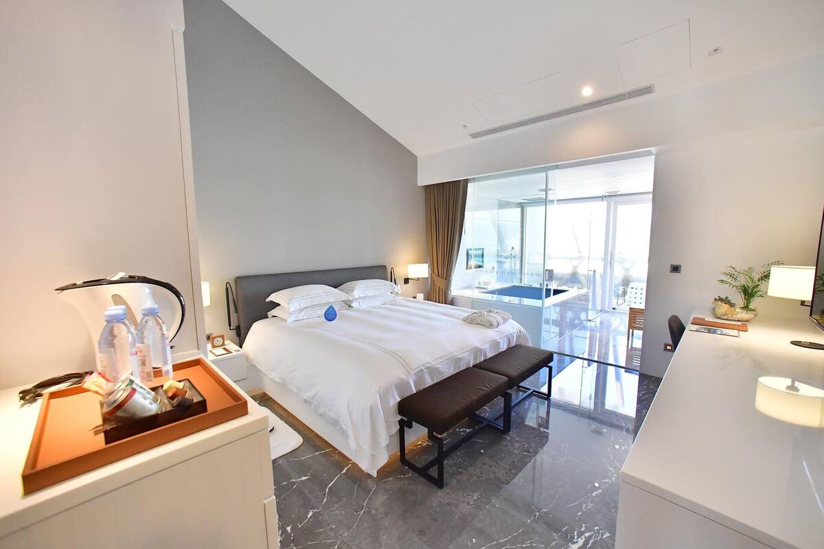 A modern hotel room with a large bed, a bench, a desk, and a view of the city through large windows.