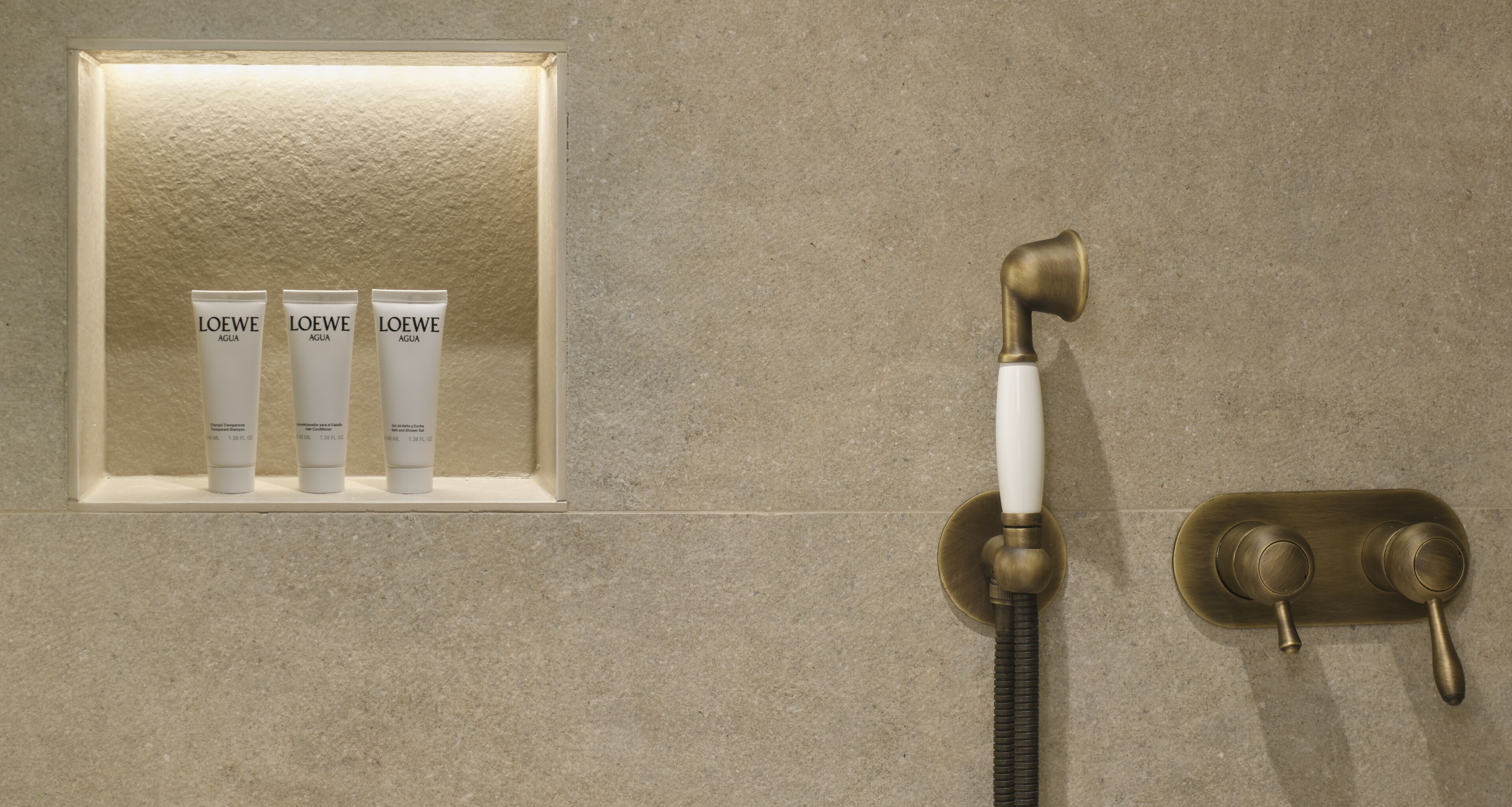 Zona de ducha con cabezal de ducha de bronce y un expositor enmarcado con tres tubos de loción etiquetados como "LOEWE AGUA".