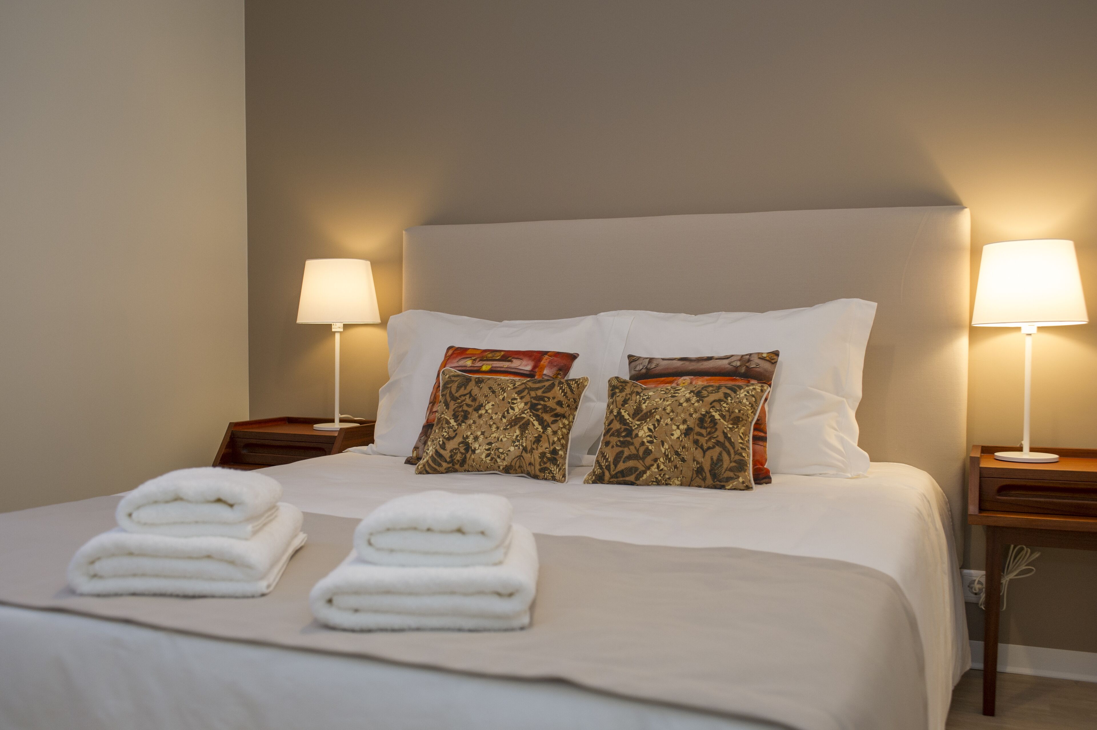Una cama bien tendida con sábanas blancas y dos almohadones decorativos, flanqueada por mesitas de noche con lámparas.
