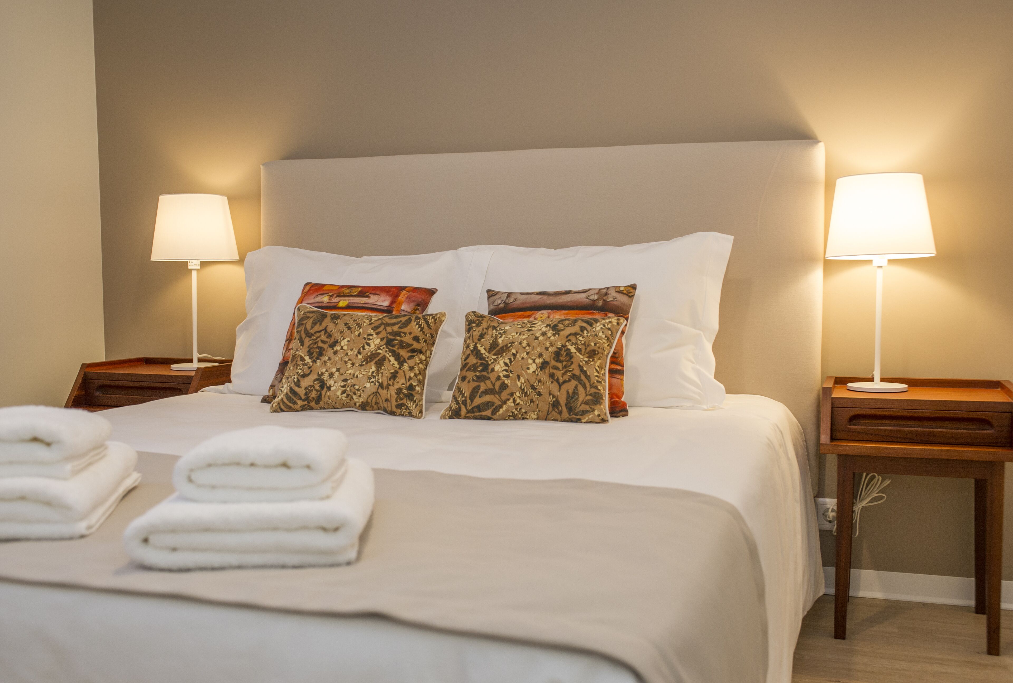Una cama bien tendida con sábanas blancas y almohadas decorativas, dos mesitas de noche con lámparas y toallas dobladas sobre la cama.