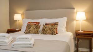 Ein ordentlich gemachtes Bett mit weißen Leinentüchern und dekorativen Kissen, zwei Nachttische mit Lampen und gefaltete Handtücher auf dem Bett.