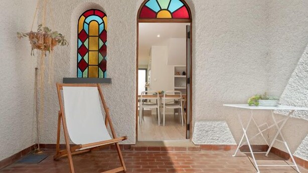 Ein Zimmer mit einer weißen, strukturierten Wand, einem hölzernen Staffelei mit einer leeren Leinwand, einem kleinen Tisch mit einer Pflanze und einem farbenfrohen Buntglasfenster.