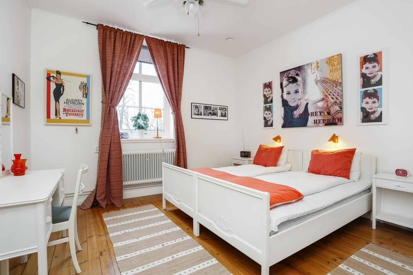 A modern bedroom with a white bed, red pillows, a desk with a chair, and a radiator.