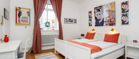 A modern bedroom with a white bed, red pillows, a desk with a chair, and a radiator.