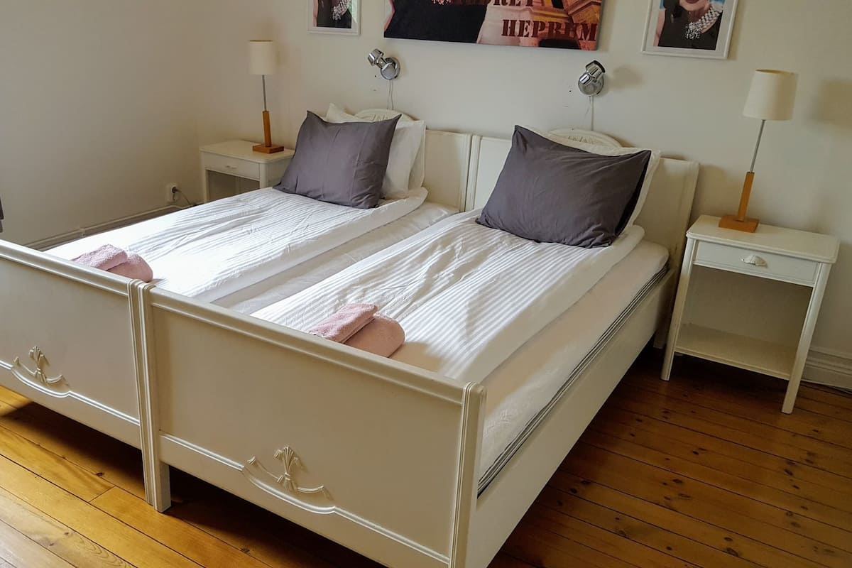 A double bed with a white headboard and footboard, two nightstands with lamps, a radiator, and a window with curtains.