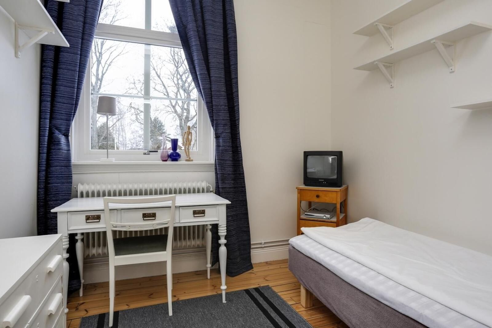 A bedroom with a single bed, a desk with a chair, a radiator, a television on a wooden stand, and a window with curtains.