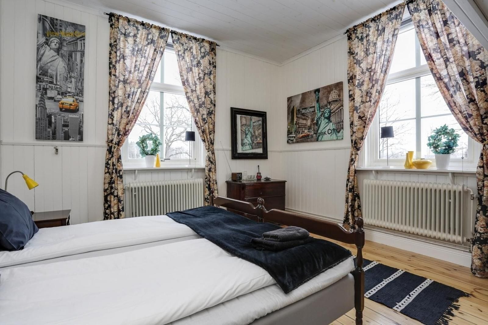 A bedroom with a bed, a dresser, a radiator, and two windows with curtains.