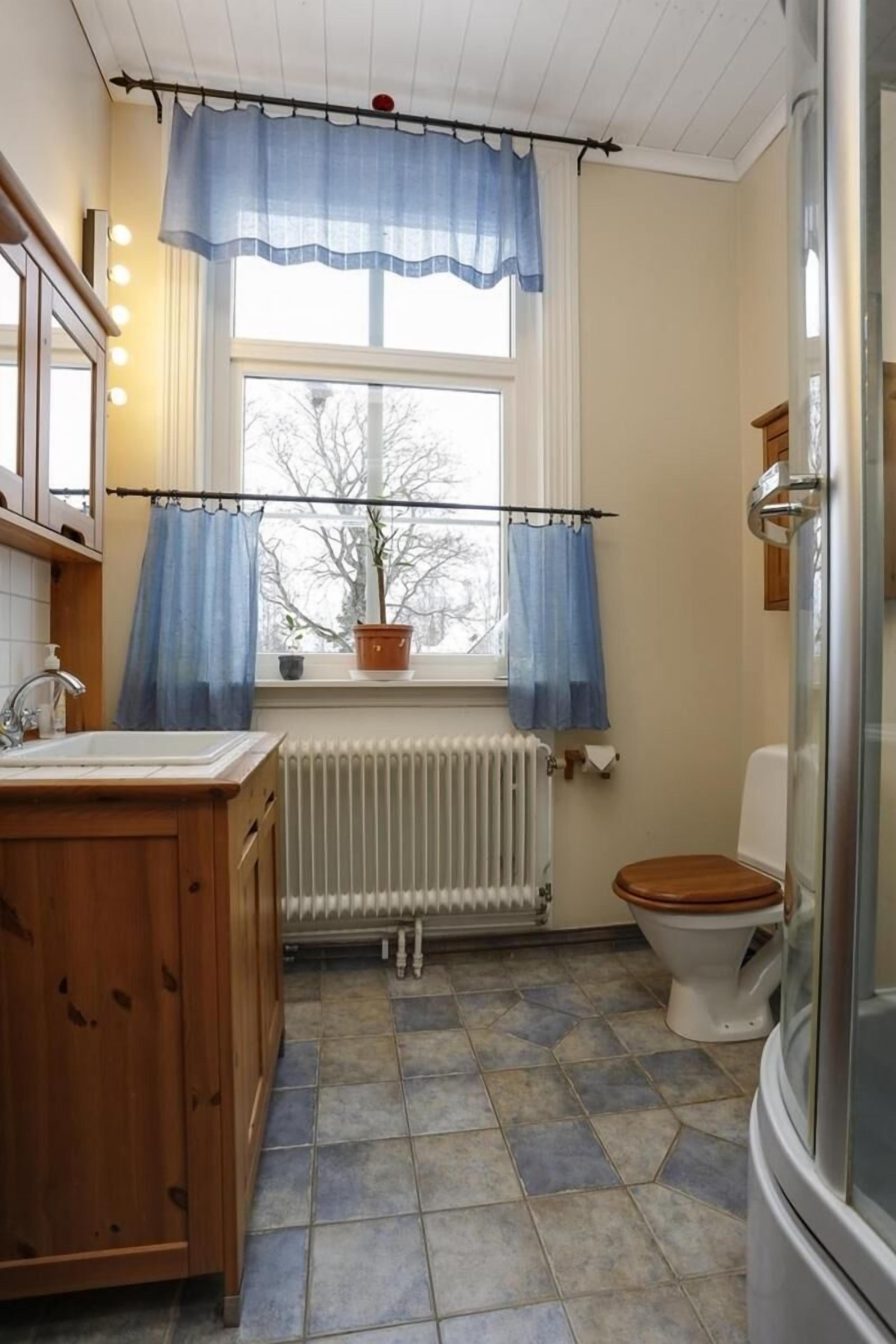 Un bany amb un tocador de fusta, un vàter blanc, una finestra amb cortines blaves i un radiador.