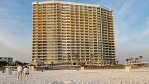 Ein Hochhaus-Hotel mit mehreren Balkonen, ein sandiger Strand im Vordergrund und Palmen im Hintergrund.