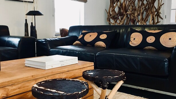 Un salon moderne avec un canapé en cuir noir, deux tabourets en bois, une table basse en bois et un grand tableau abstrait accroché au mur.