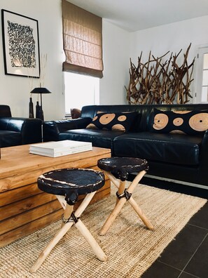 A modern living room with a black leather sofa, two wooden stools, a wooden coffee table, and a large abstract painting on the wall.