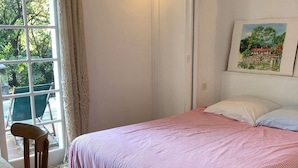 Een slaapkamer met een bed, een stoel, een tafel, een raam met gordijnen, een wandlamp en een lijst met een afbeelding.