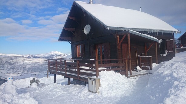 Una cabaña de madera con techo nevado y una terraza de madera, rodeada por un paisaje invernal y montañas al fondo.