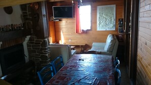 Una habitación acogedora con chimenea de piedra, un televisor montado en la pared, una mesa de madera con mantel de plástico y una zona de estar con cojines.