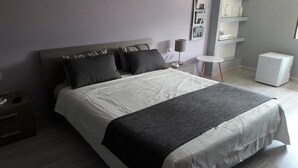 A neatly made bed with a grey blanket, a bedside table with a lamp, and a small white refrigerator in a modern hotel room. - Campo Sportivo (Taurianova)