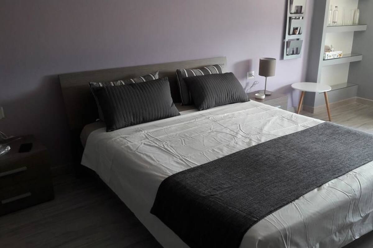 Ein ordentlich bezogenes Bett mit grauer Decke, ein Nachttisch mit Lampe und ein kleiner, weißer Kühlschrank in einem modernen Hotelzimmer.
