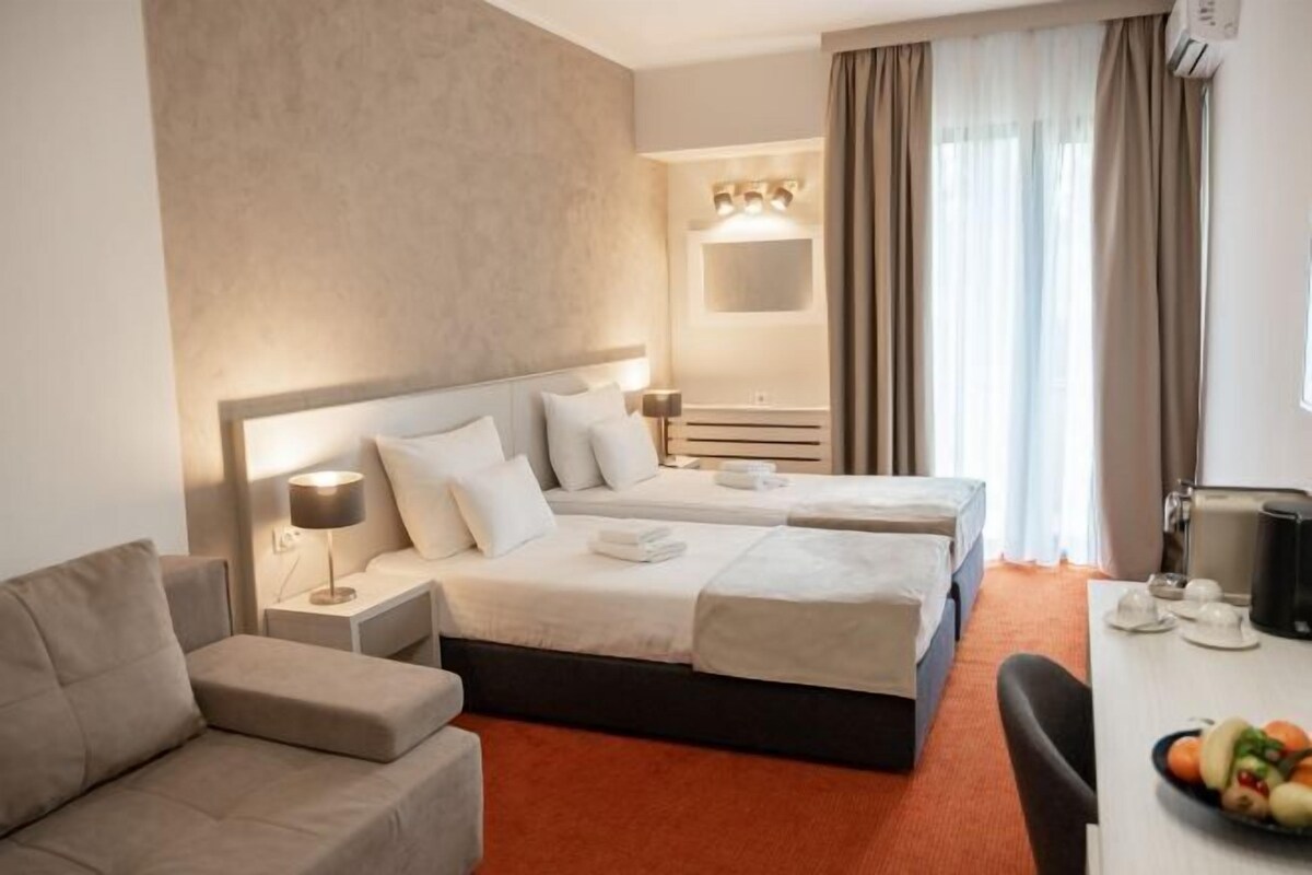 Quarto de hotel com duas camas, um sofá, uma escrivaninha com uma tigela de frutas e um espelho sobre a cama.