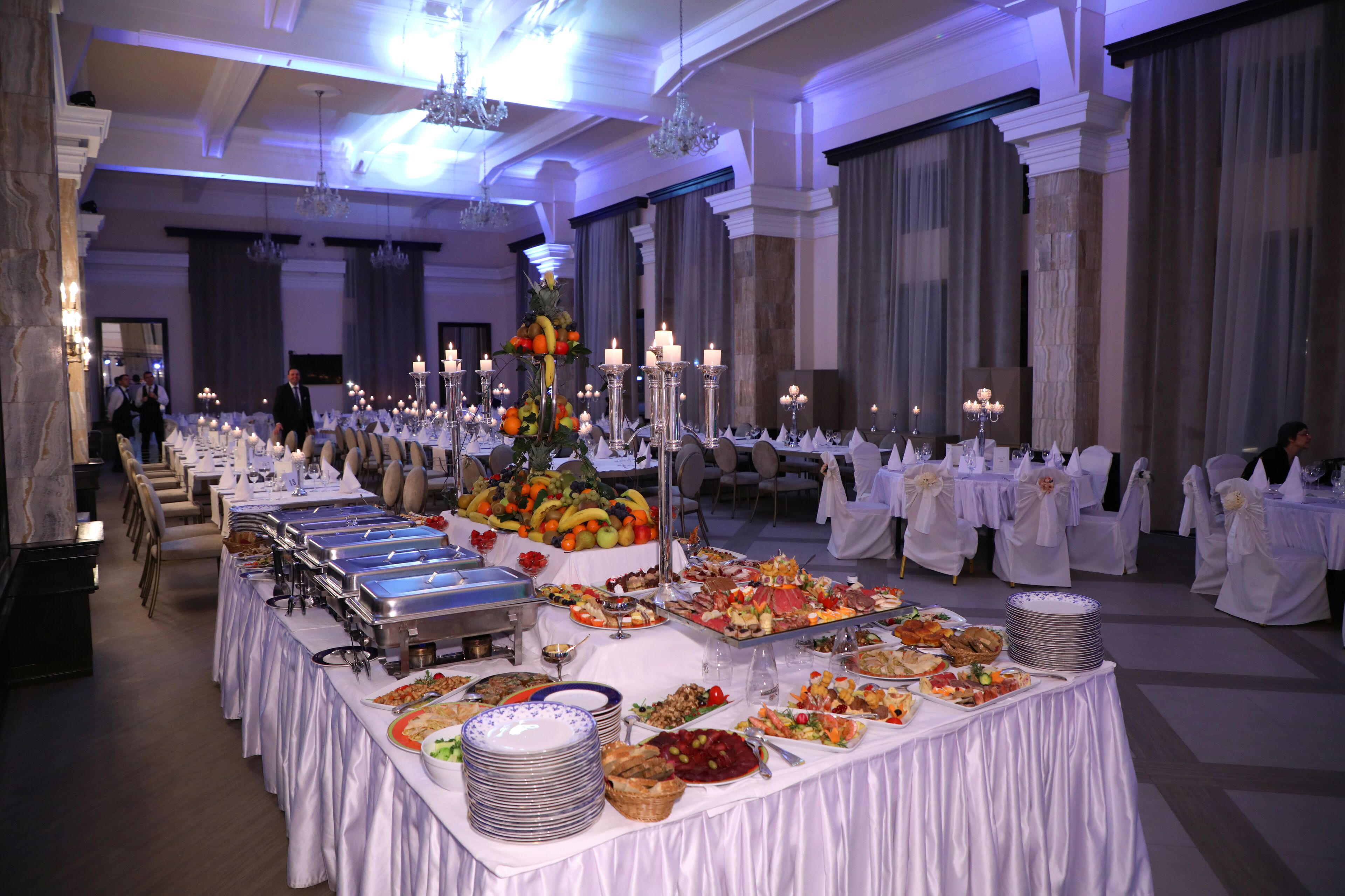 A banquet hall with multiple buffet tables covered in white tablecloths, featuring a variety of food items and candles.