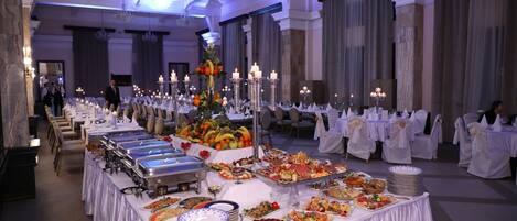 A banquet hall with multiple buffet tables covered in white tablecloths, featuring a variety of food items and candles.