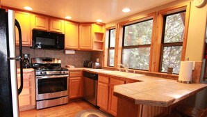 Una cocina con armarios de madera, electrodomésticos de acero inoxidable, microondas integrado y un amplio ventanal con vistas a árboles.