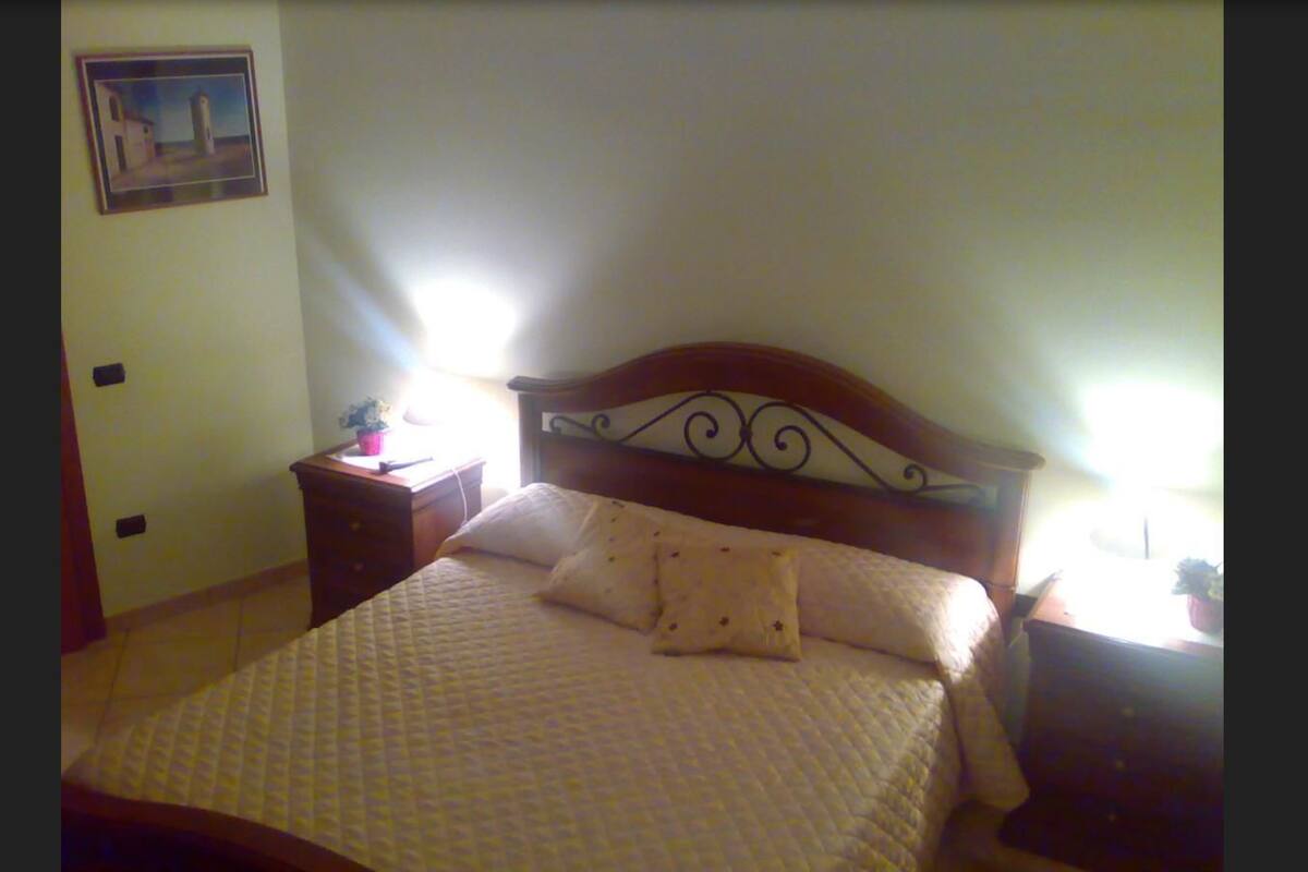A neatly made bed with a quilted cover, a wooden headboard with a decorative design, a bedside table with a lamp, and a framed picture on the wall.