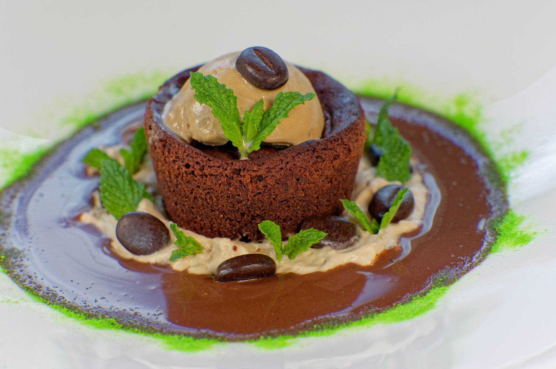Dessert med chokoladebund, en isbold og en kaffebønne ovenpå, omgivet af mynteblade og chokoladestykker.
