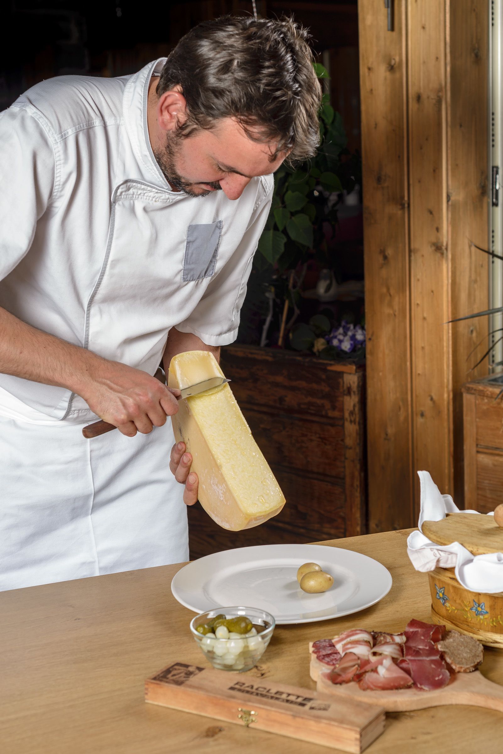 Una persona corta un bloque grande de queso sobre una mesa de madera, con un plato y un bol de aceitunas, además de una tabla de madera con fiambres.