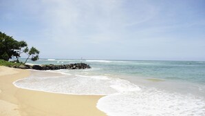 Ein sandiger Strand mit leichten Wellen, einem Felsen und einem wolkenlosen Himmel.