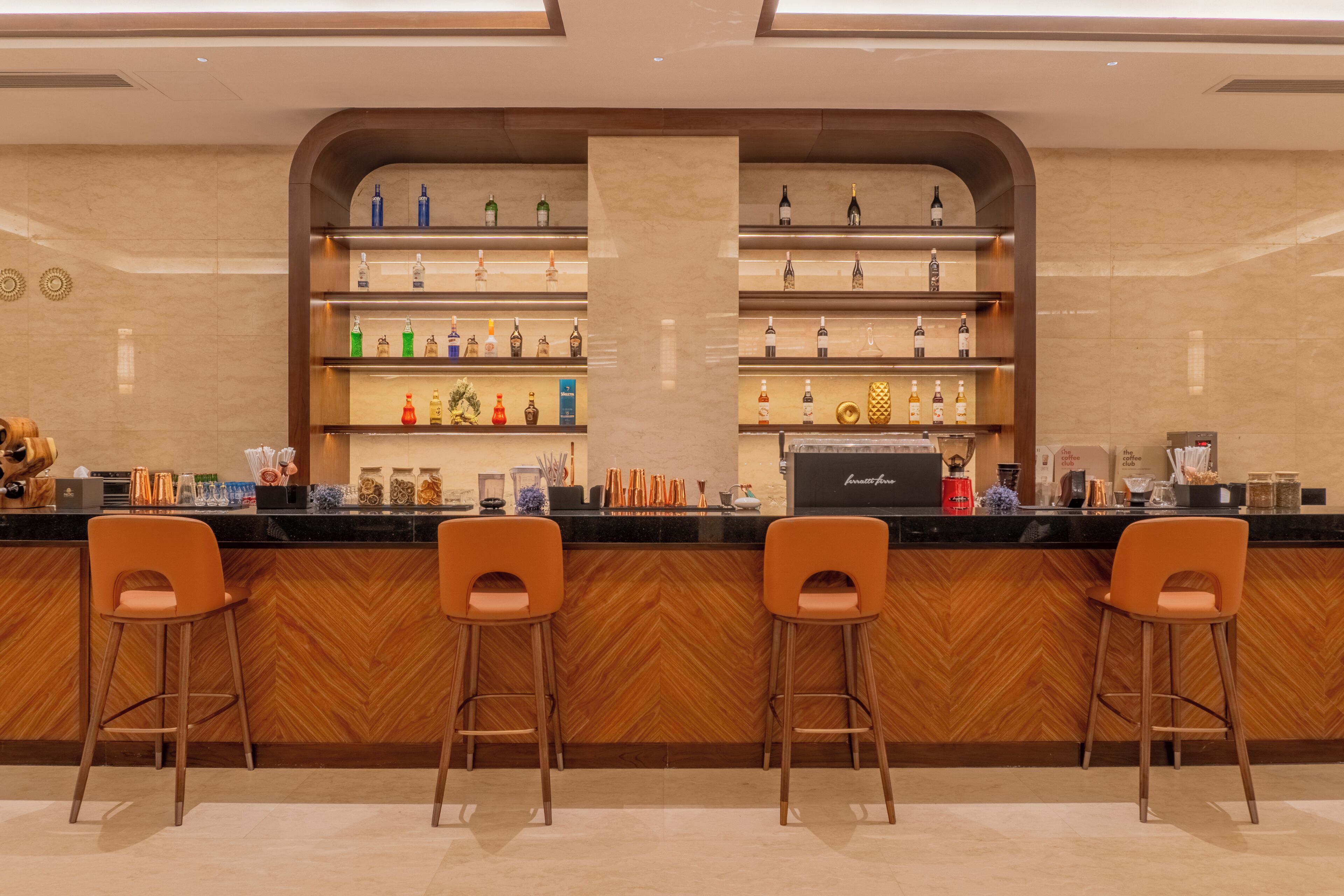 A bar area with a wooden herringbone floor, two orange bar stools, and shelves stocked with various bottles and bar tools.