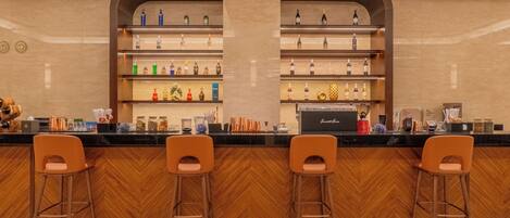 Uma área de bar com chão de madeira em padrão herringbone, dois banquetas laranja e prateleiras repletas de garrafas e utensílios de bar.