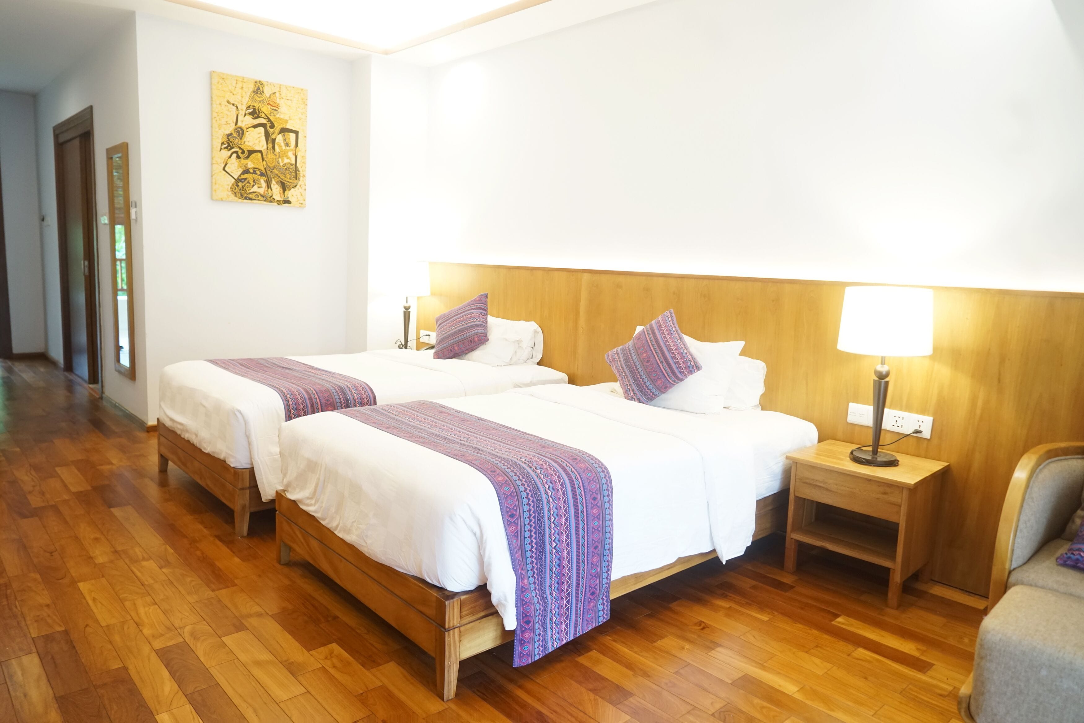 Een hotelkamer met twee bedden, houten hoofdeinden, een nachtkastje met een lamp en een schilderij aan de muur.