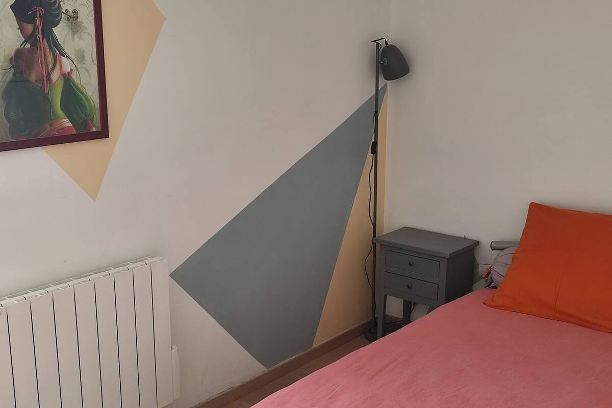 A bedroom with a bed, a nightstand, a floor lamp, a radiator, and a painting on the wall.
