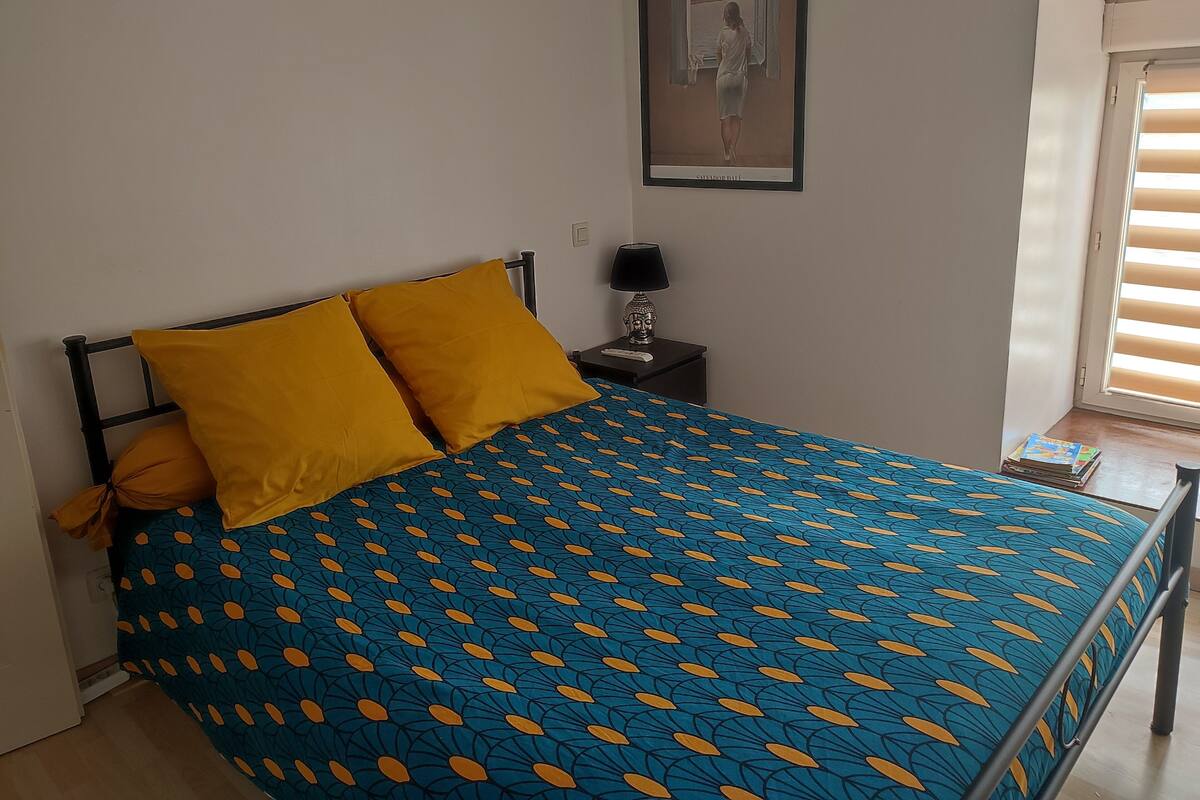 A bed with a blue and yellow patterned bedspread, a nightstand with a lamp, and a window with blinds.