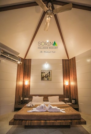 A hotel room with a large bed, wooden paneling, two wall-mounted lights, and a ceiling fan. - Sorina Hillside Resort (Pune)