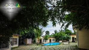 A pool area with a tiled deck, surrounded by lush greenery and a clear blue sky. - Sorina Hillside Resort (Pune)