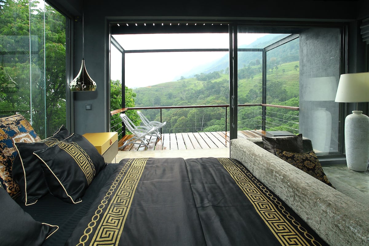 A bedroom with a large window, a bed with patterned bedding, a chair, a table, and a lamp.