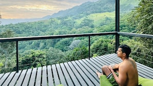 A man sitting on a wooden deck with a green cushion, overlooking a lush green landscape with hills and a cloudy sky. - Romance Valley (Hildummulla)