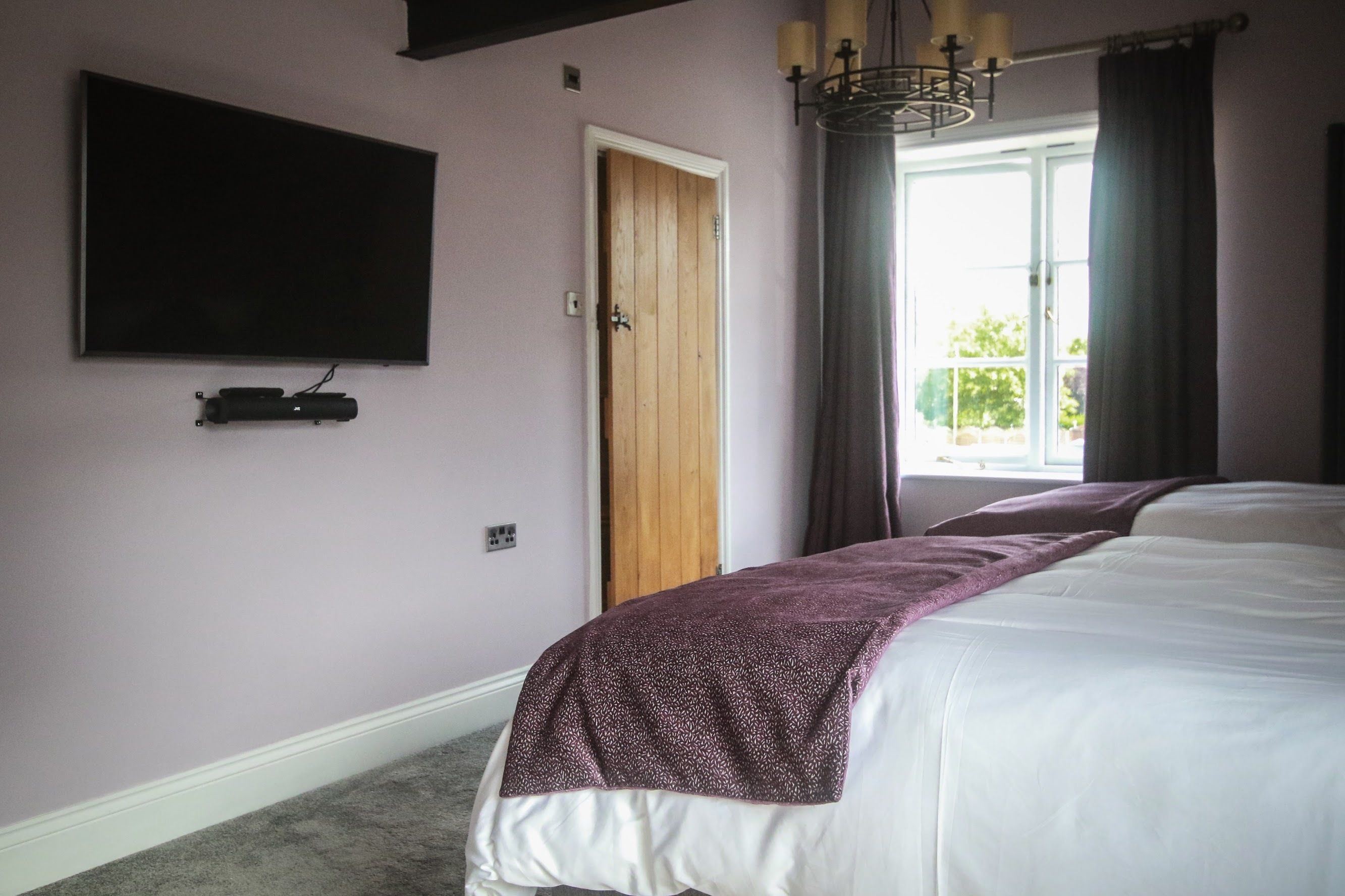 A bedroom with a large bed, a flat-screen TV mounted on the wall, a wooden door, and a window with curtains.