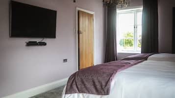 A bedroom with a large bed, a flat-screen TV mounted on the wall, a wooden door, and a window with curtains.