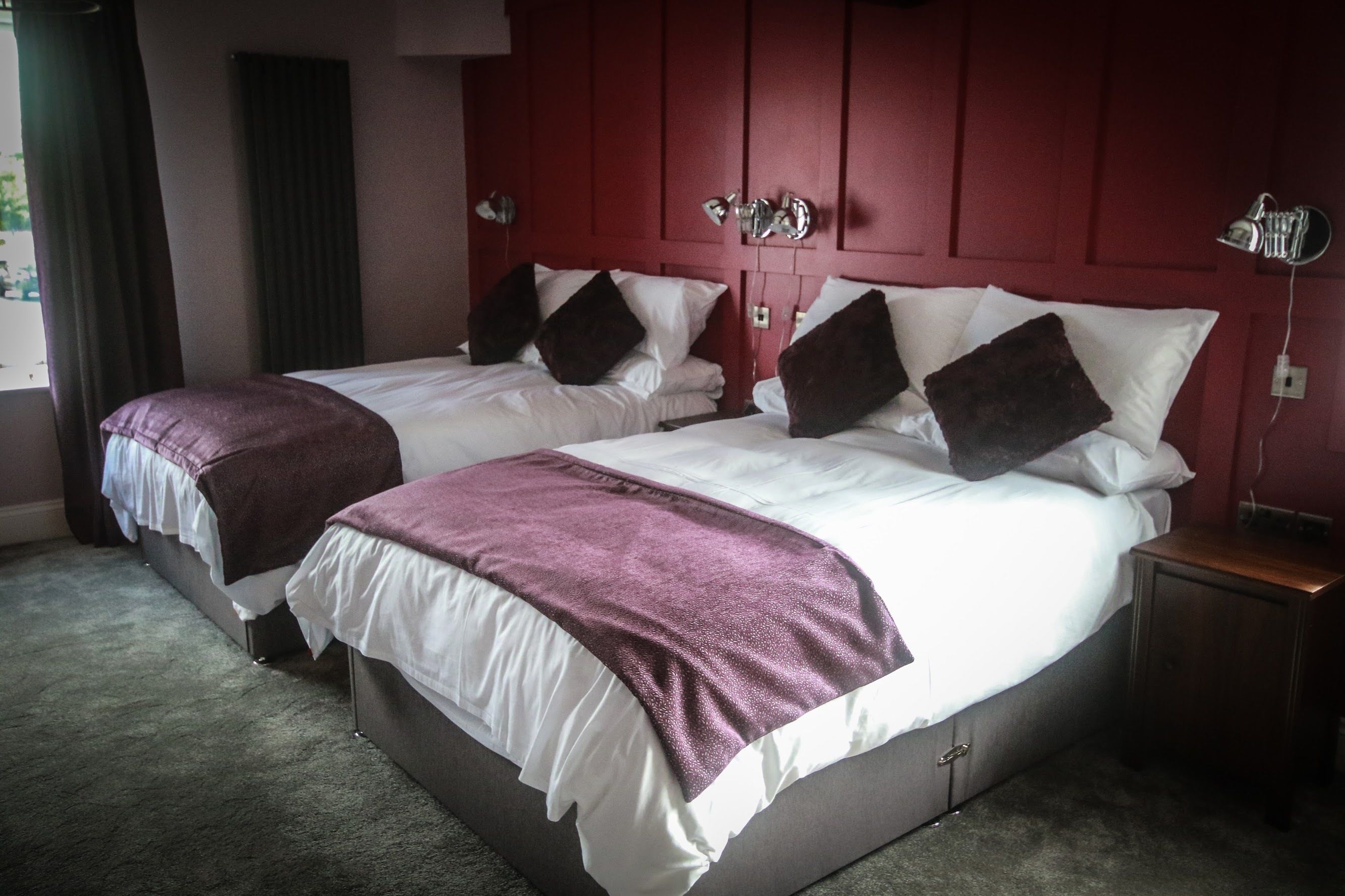 Habitación de hotel con dos camas, cada una con sábanas blancas y almohadas oscuras. Tiene un cabecero rojo, una lámpara de noche y una mesita de noche de madera.