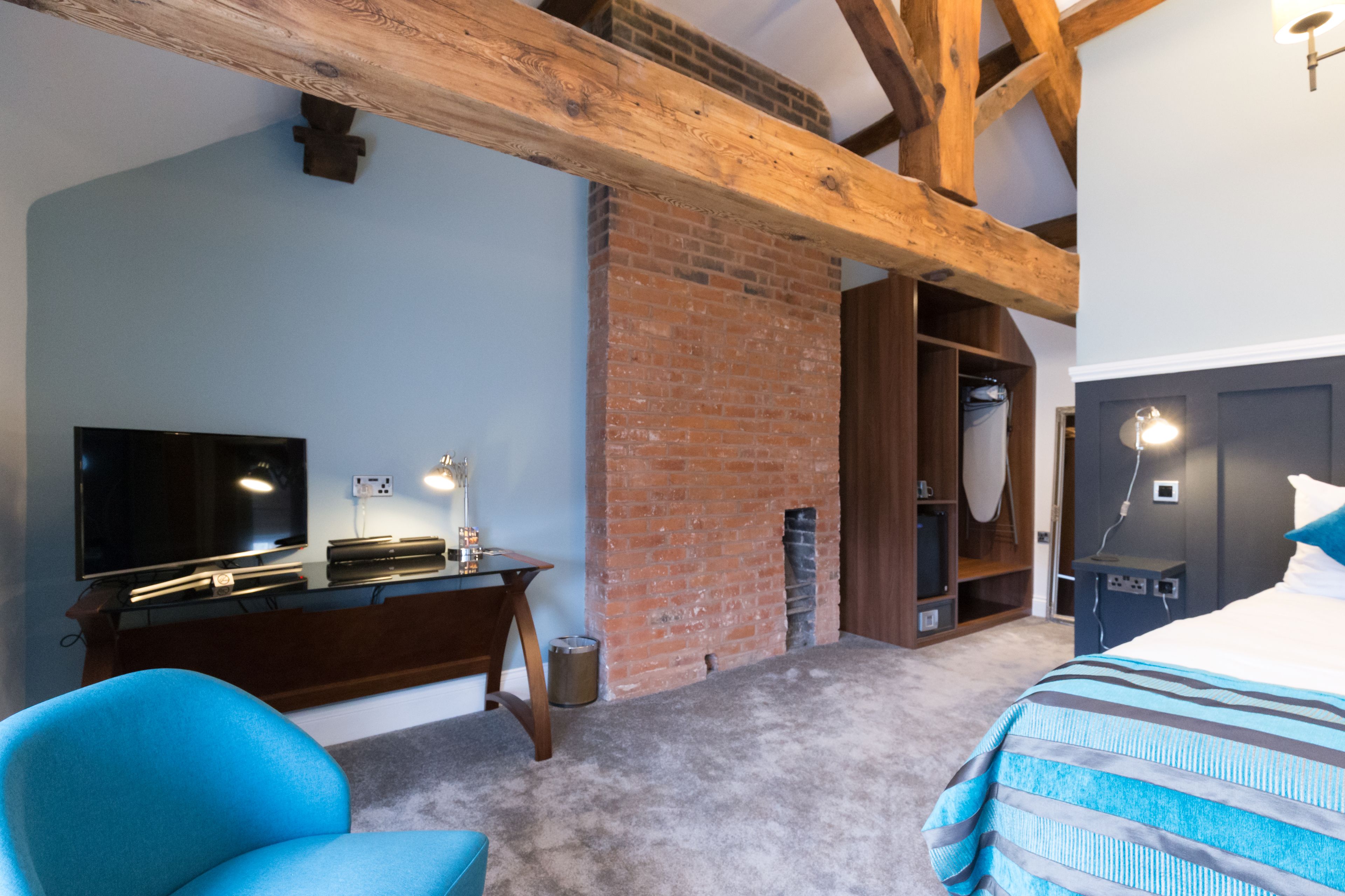 Ein Schlafzimmer mit freiliegenden Holzbalken, einem Kamin aus Backsteinen, einem Flachbildfernseher, einem Schreibtisch und einem Bett mit gestreifter Bettwäsche.