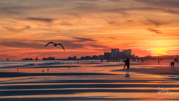 Une scène de plage au coucher du soleil, avec des personnes qui se promènent et, en arrière-plan, la silhouette d’une ville.