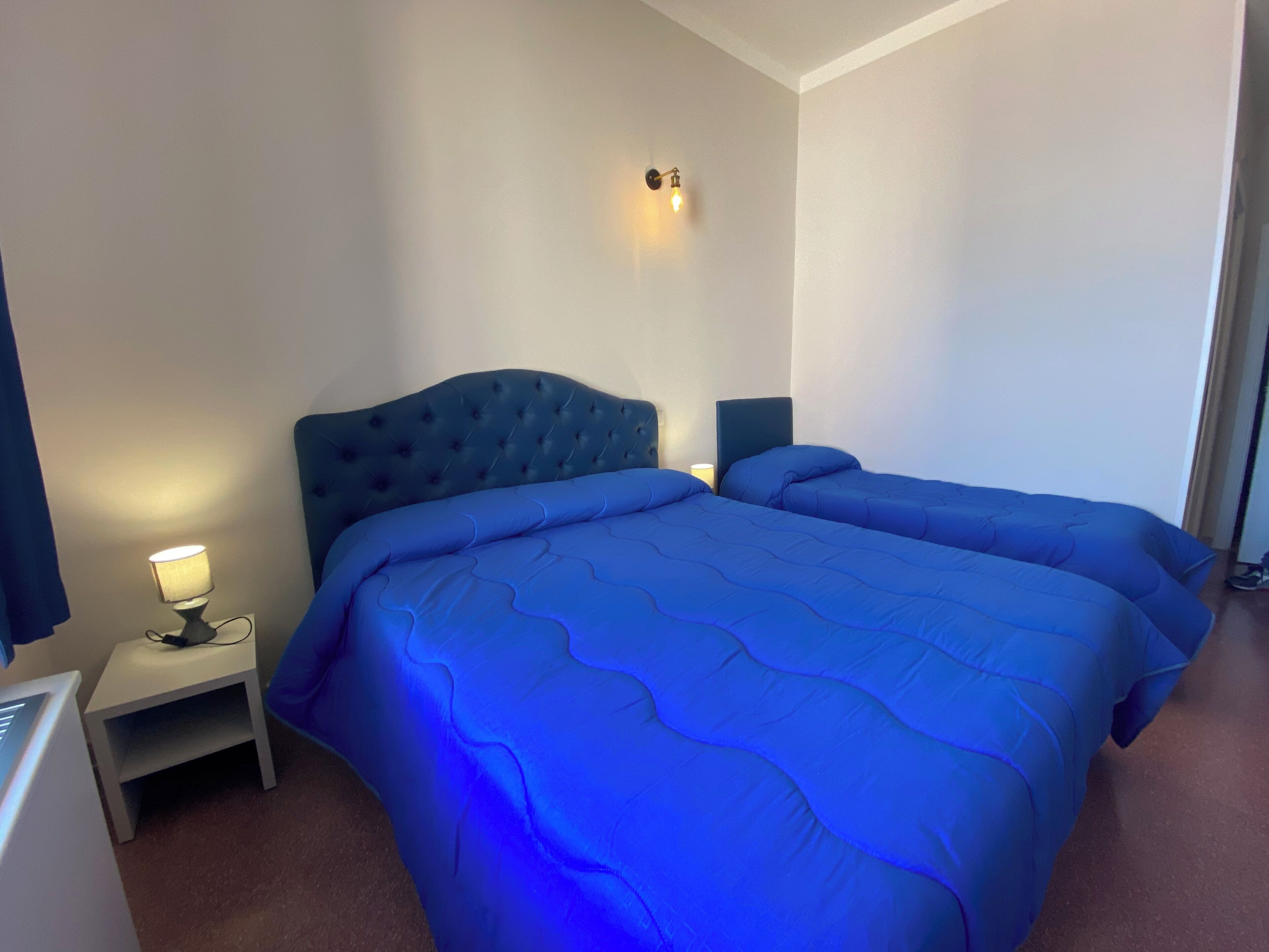 A bedroom with two beds, a nightstand, a lamp, and a wall-mounted light fixture.