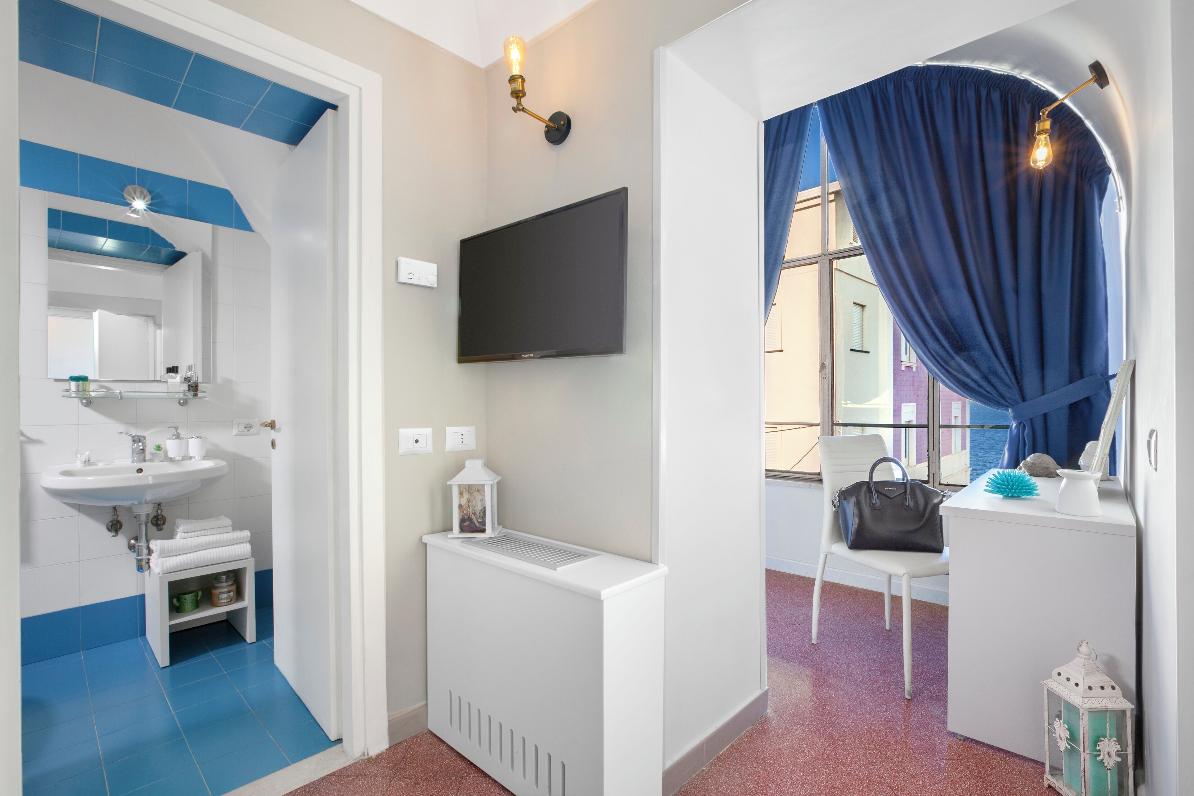 Une salle de bain moderne avec un lavabo, un miroir et un radiateur. Un téléviseur est fixé au mur, et une chaise avec un sac à main se trouve près d’une fenêtre ornée de rideaux bleus.