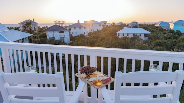 Een balkon met een dienblad eten en twee witte stoelen, met uitzicht op een kustgebied met gebouwen en bomen tijdens zonsondergang.