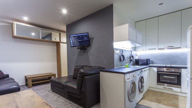 A compact living space with a sofa, a washing machine, a small kitchen area, and a mounted TV. - Penmoor Chance - Old Town (Bratislava)
