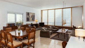 Un salón espacioso con zona de comedor, ventanas grandes con persianas, televisor de pantalla plana y chimenea.