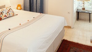 Un dormitorio con cama, mesita de noche, lámpara, ventana con cortinas y puerta que da a otra habitación.
